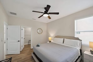 A bedroom with a bed, a ceiling fan, a bedside table with a lamp, a window with blinds, and a door leading to another room.