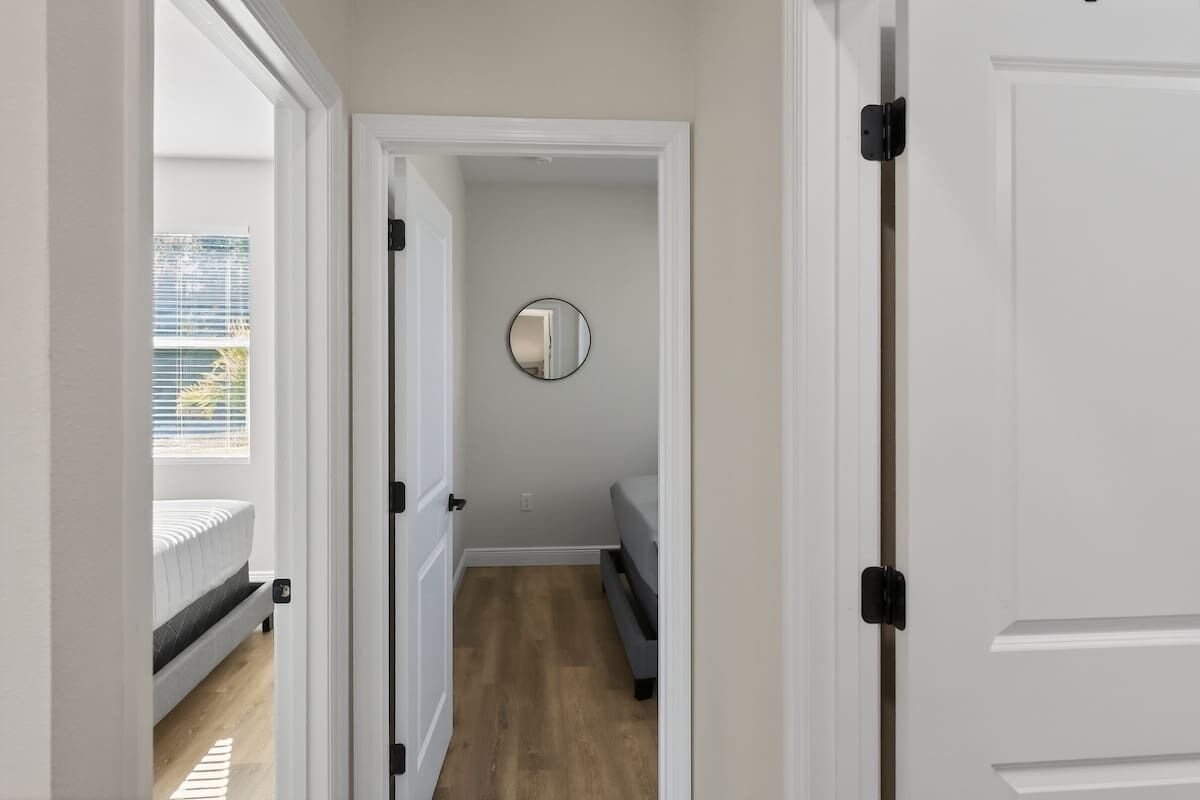 Een smalle gang met een bed aan de linkerkant, een ronde spiegel aan de muur en een deur aan de rechterkant.