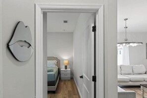 A hallway leading to a bedroom with a bed, bedside table, and a lamp. There is a white door on the right and a wall-mounted light fixture on the left. - Beautiful 4/2 Oasis: Beaches Nearby | w/ King Bed (Englewood)