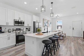 A modern kitchen with white cabinets, stainless steel appliances, a central island with stools, pendant lighting, and artwork on the walls.