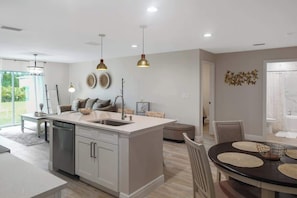 A modern kitchen with a central island, pendant lighting, and a dining area with a round table. - Coastal Escape: New Luxury House (Port Charlotte)