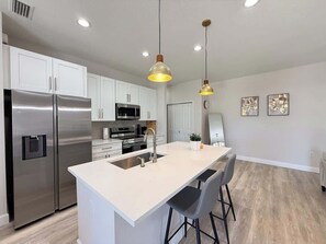 Een moderne keuken met roestvrijstalen apparatuur, een centraal eiland met barkrukken, hanglampen en uitzicht op de woonkamer.
