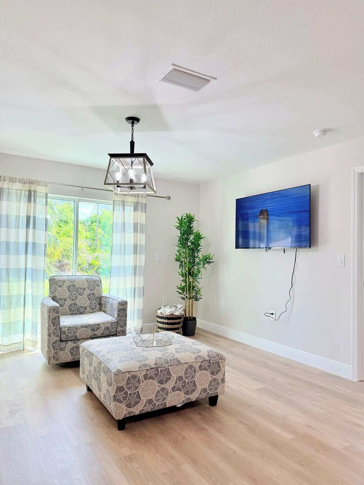 Un salón amplio con un sofá moderno, un reposapiés estampado, un televisor de pantalla plana grande montado en la pared y una planta en maceta.