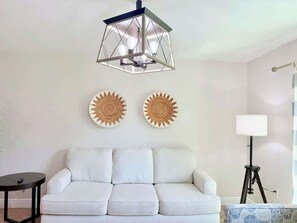 Ein weißes Sofa mit einer geometrischen Pendelleuchte darüber, zwei gewebte Wanddekorationen, eine Stehlampe und ein kleiner Tisch.