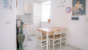 A kitchen with white cabinets, a wooden table, and chairs. There is a window with a view of the outside, a clock on the wall, and a vase on the counter.