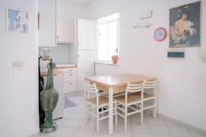 A kitchen with white cabinets, a wooden table, and chairs. There is a window with a view of the outside, a clock on the wall, and a vase on the counter.