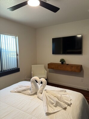 A hotel room with a bed, a ceiling fan, a mounted TV, a chair, and a window with blinds. - Cozy 3-Bedroom Apartment | 15 Minutes from The Strip (Las Vegas)