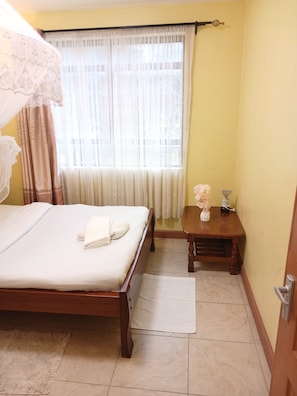A hotel room with a bed, a bedside table, a chair, a window with curtains, and a small table with a vase.