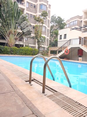 A swimming pool with metal railings, surrounded by greenery and a multi-story building with balconies.
