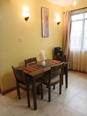 A dining area with a wooden table, chairs, a vase with flowers, a coffee maker, and a painting on the wall. - The Terminal Residence (JWM9+X5X, Nairobi, Kenya)
