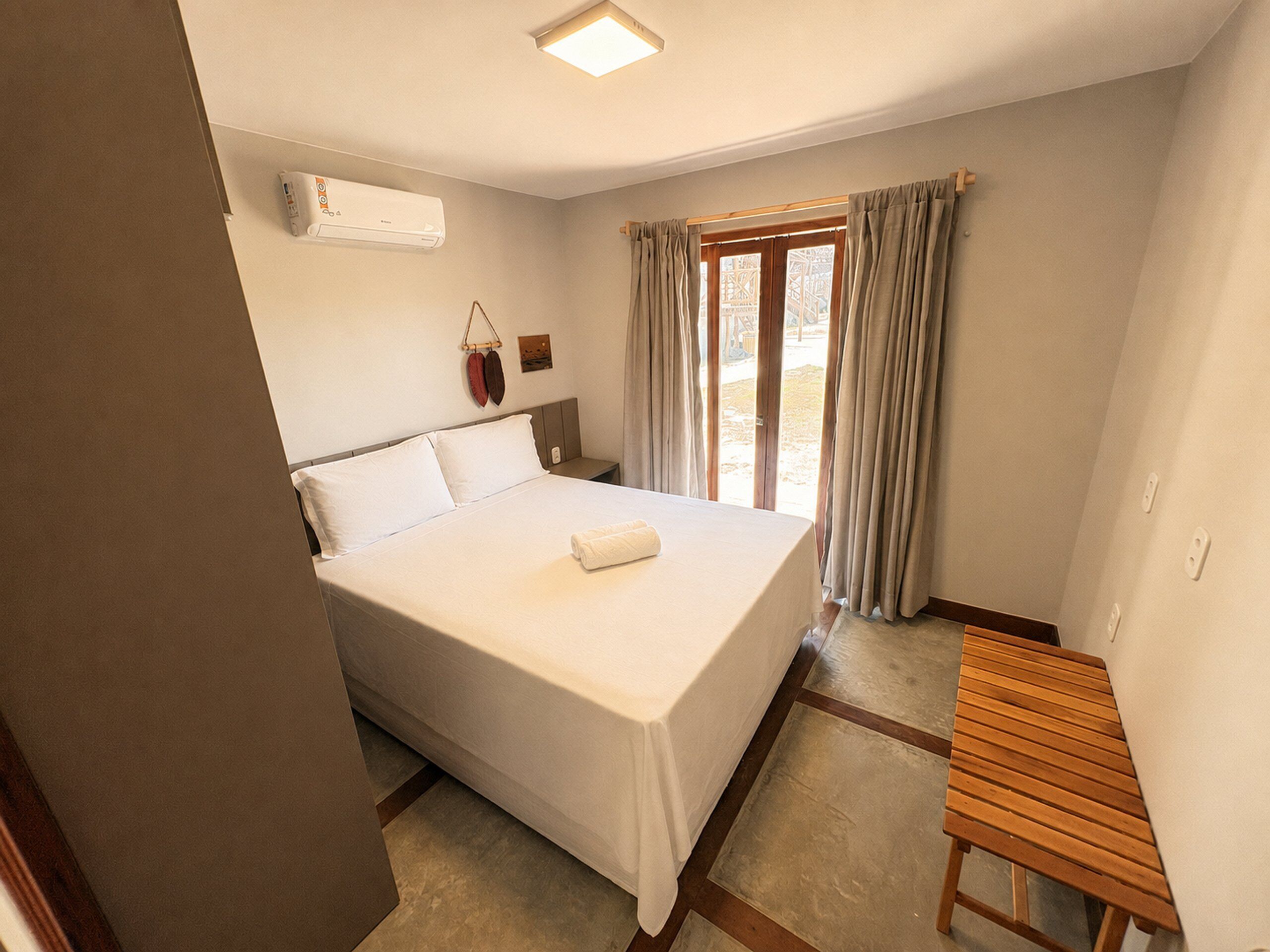 A hotel room with a bed, a bedside table, a window with curtains, and an air conditioning unit.