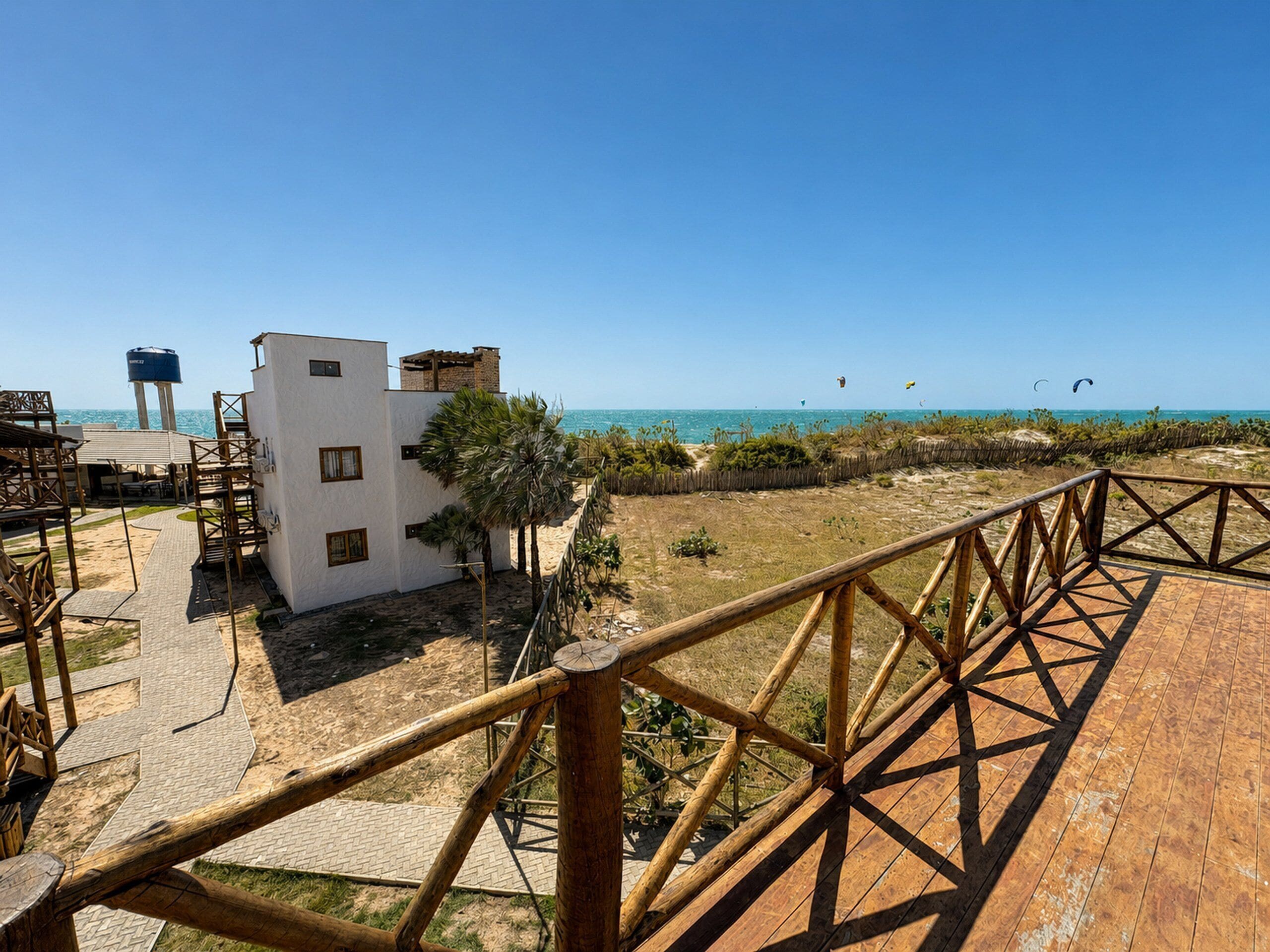Um deck de madeira com guarda-corpo com vista para uma praia, um edifício branco com várias janelas e um céu azul claro.