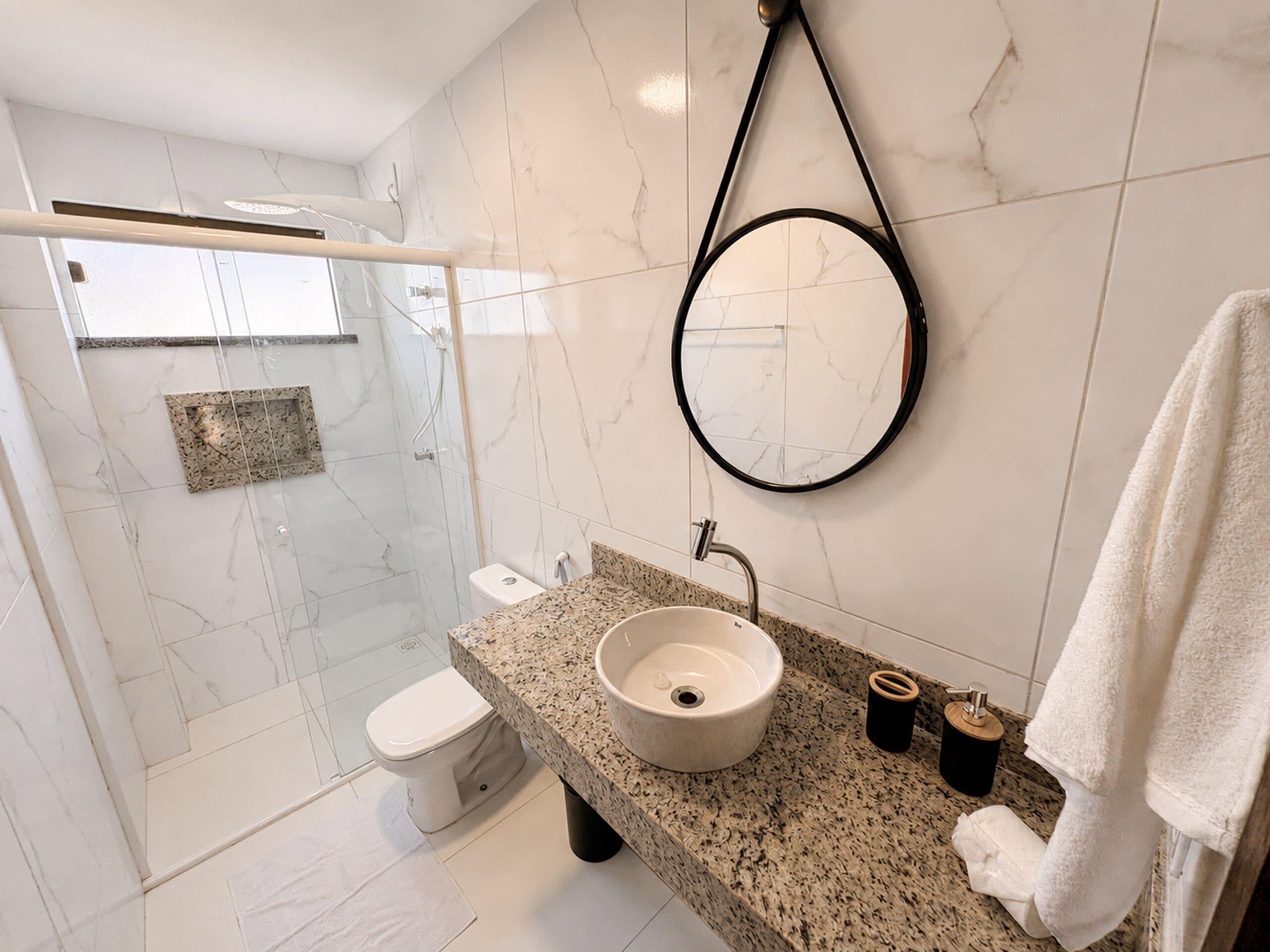 A modern bathroom with a glass-enclosed shower, a white toilet, a granite countertop with a sink, and a round mirror.