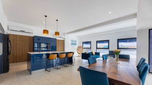 Een moderne keuken met een centraal eiland, barkrukken en een eethoek met een houten tafel en blauwe stoelen.