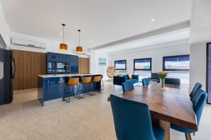 A modern kitchen with a central island, bar stools, and a dining area with a wooden table and blue chairs. - Handelskade's Grand Historical Penthouse: Refined Luxury Living (Willemstad)