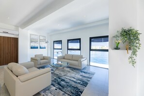 A spacious living room with a large window overlooking a body of water, a modern sofa set, and a glass coffee table. - Handelskade's Grand Historical Penthouse: Refined Luxury Living (Willemstad)