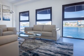 A modern living room with a glass coffee table, beige sofa, and large windows overlooking a body of water. - Handelskade's Grand Historical Penthouse: Refined Luxury Living (Willemstad)