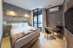 A hotel room with a bed, a desk, and a chair. There is a window with a view of buildings. - Address by Bhomy (São Paulo)