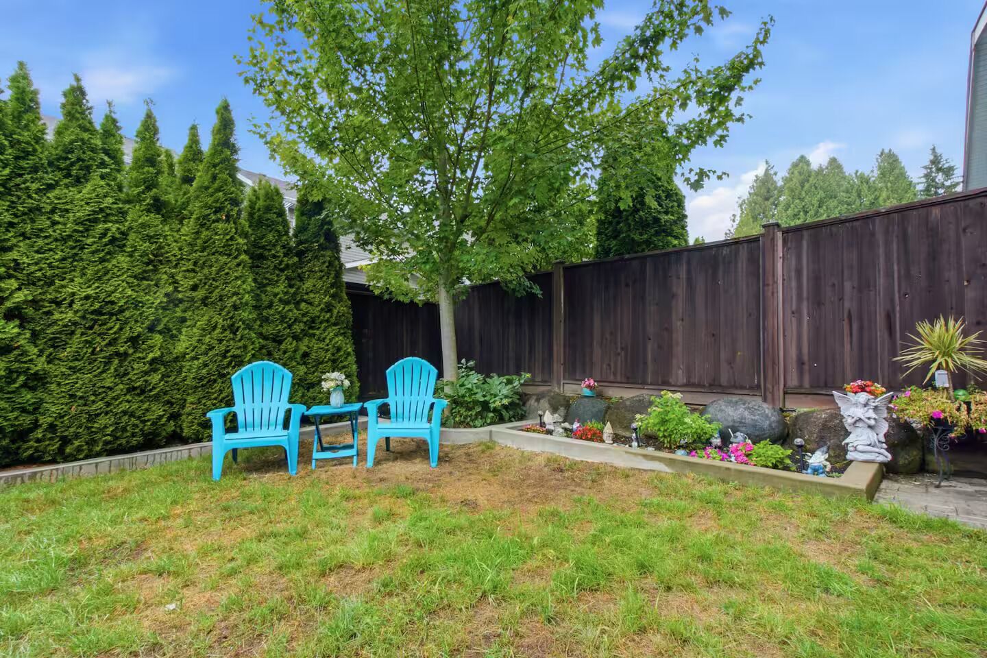 A backyard with a wooden fence, a tree, a small garden with various plants, and two blue chairs.