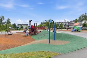 A playground with various play structures, including slides and climbing areas, surrounded by a landscaped area with mulch and grass.