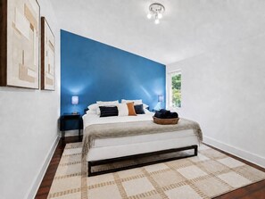A bedroom with a bed, bedside tables, a nightstand, a window, and a wall with a blue accent.