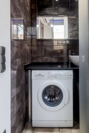 Un baño moderno con baldosas oscuras, una lavadora blanca y un espejo que refleja la habitación.