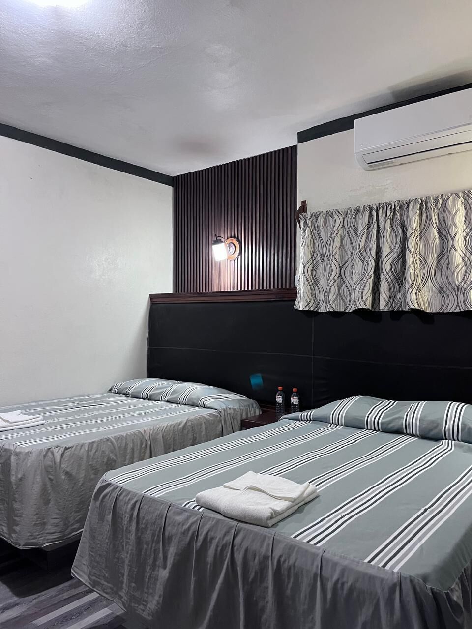 Quarto de hotel com duas camas, ar-condicionado fixado na parede e janela com cortinas estampadas.