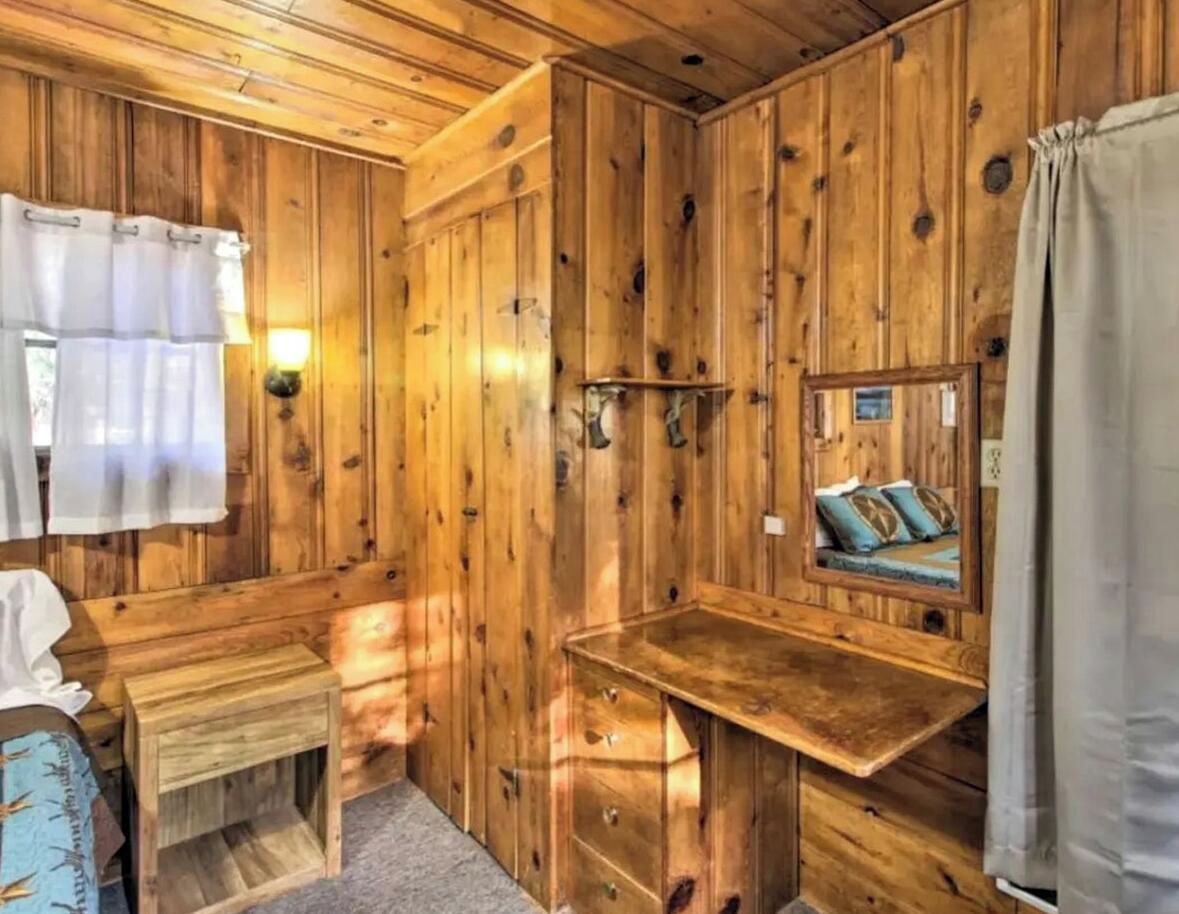 A wooden cabin interior with a bed, a small table, a mirror, and a window with curtains.