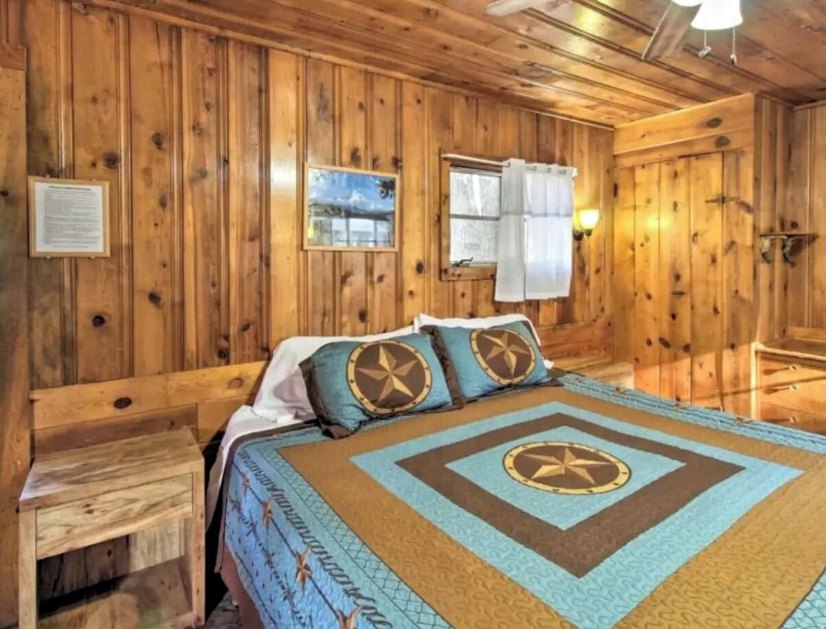 A wooden cabin bedroom with a bed, a nightstand, a window with curtains, and a wall-mounted light fixture.