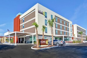 Ein mehrstöckiges Hotel mit modernem Design, das einen überdachten Eingangsbereich mit Palmen und ein Schild mit der Aufschrift Home 2 Suites by Hilton zeigt.
