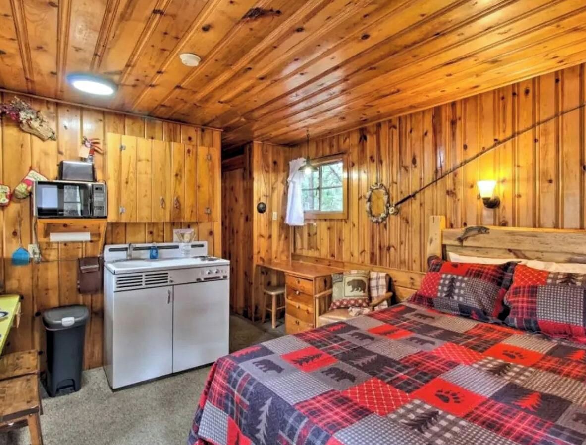 A cabin-style room with a wooden interior, a bed with a patterned blanket, a kitchenette with a stove, and a window.