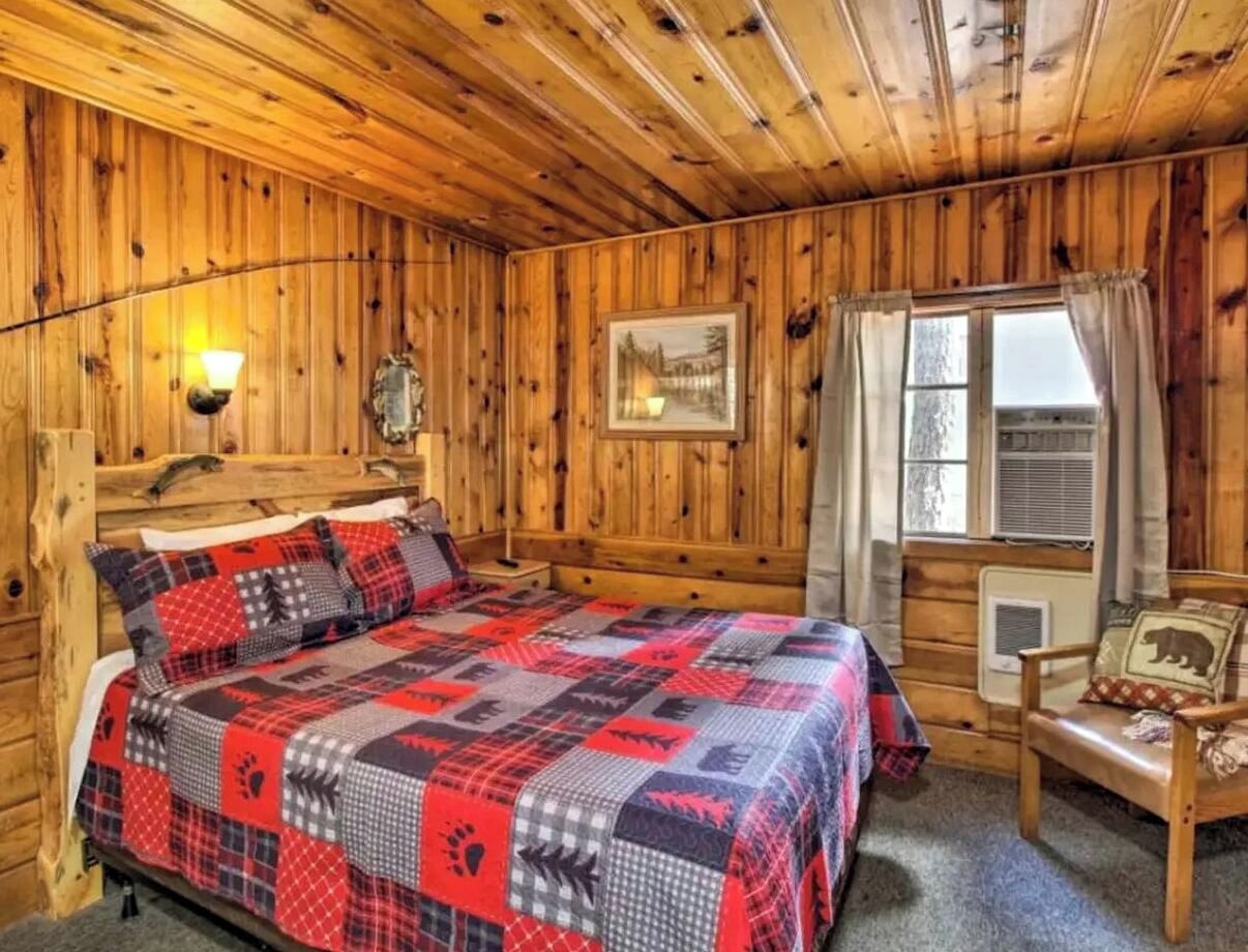 L’intérieur d’un chalet en bois, avec un lit, une chaise, une lampe fixée au mur et une fenêtre avec des rideaux.