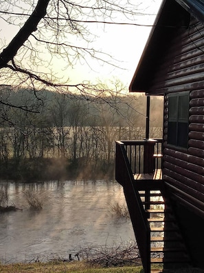 A cabin with a deck overlooking a body of water, surrounded by trees. - Riverfront Blue Heron Cabin on the Kings River (Berryville)