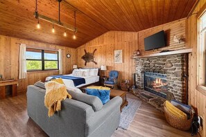 A cozy room with a bed, a fireplace, a television, and a large window. - Riverfront Blue Heron Cabin on the Kings River (Berryville)