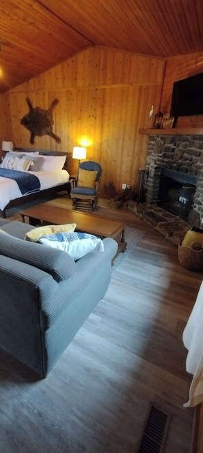 A cozy room with a bed, a sofa, a chair, a fireplace, and a wooden ceiling. - Riverfront Blue Heron Cabin on the Kings River (Berryville)