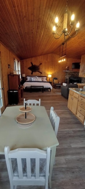 A wooden interior with a dining table, chairs, a bed, a fireplace, and a chandelier. - Riverfront Blue Heron Cabin on the Kings River (Berryville)