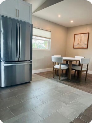 A modern kitchen with a stainless steel refrigerator, a dining table with chairs, and a window with blinds. - The Wells (Fort Myers Beach)