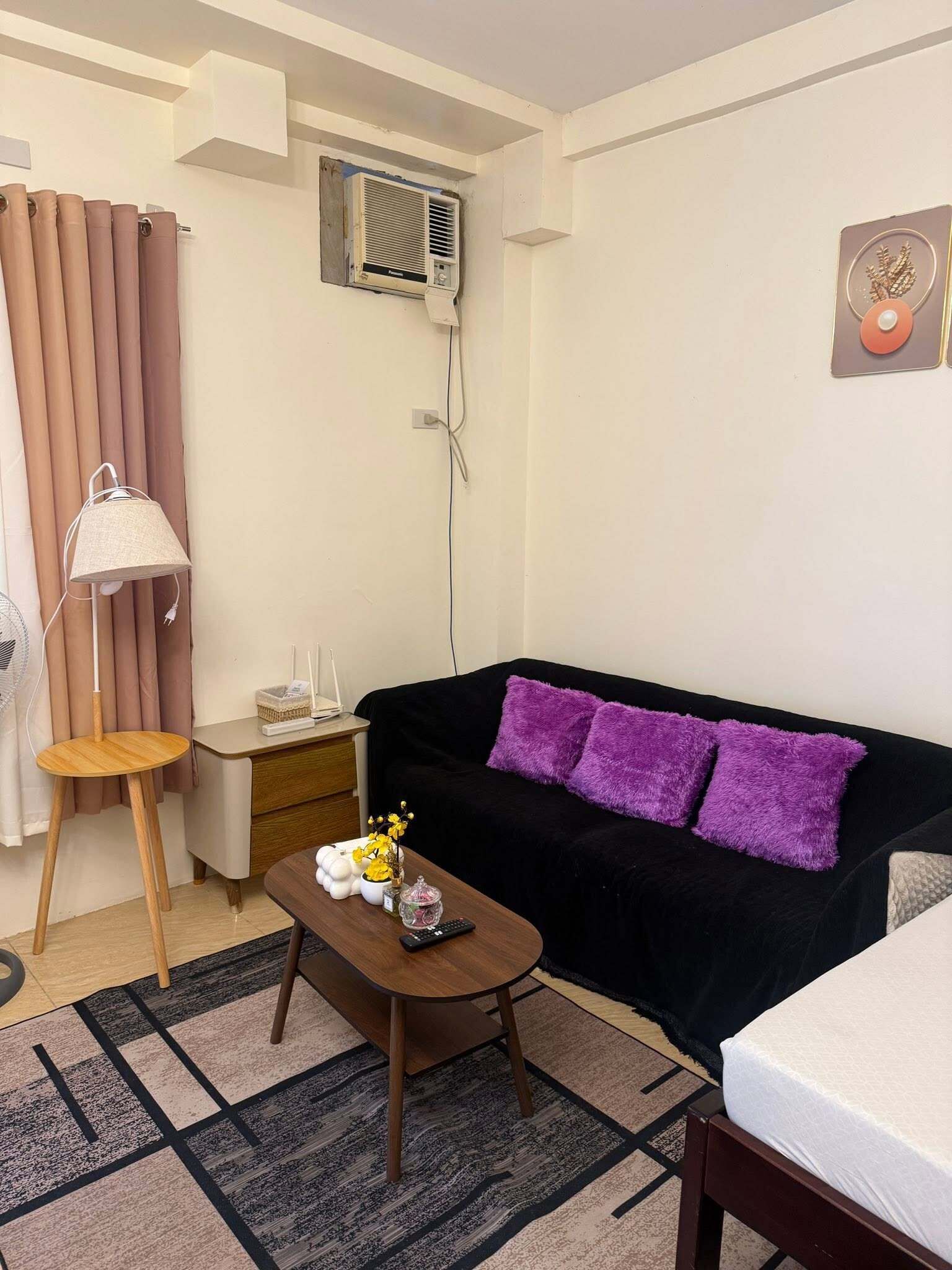 A living room with a black sofa, a wooden coffee table, a lamp, a small side table, and a wall-mounted air conditioner.