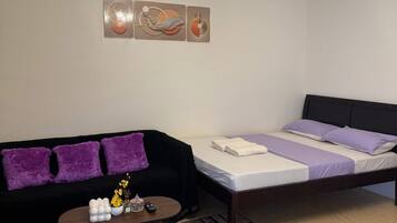 A room with a black sofa, a bed with purple bedding, a coffee table with decorative items, and a patterned rug.