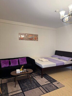 A room with a black sofa, a bed with purple bedding, a coffee table with decorative items, and a patterned rug. - Boracay Affordable Stay (Malay)