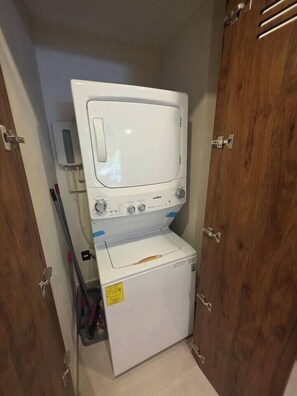 A compact laundry room with a washing machine and dryer, wooden doors, and a small shelf with cleaning supplies. - Condo 2 recámaras a pasos de playa Santa Cruz (Bahía de Santa Cruz Huatulco)