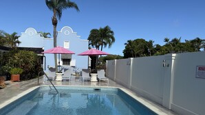 A swimming pool with lounge chairs and umbrellas, surrounded by a white wall and palm trees.