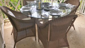 A round glass table with wicker chairs on a balcony with a white railing, surrounded by greenery.