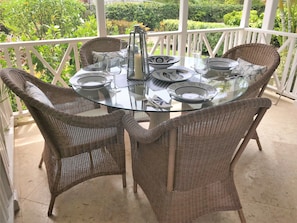A round glass table with wicker chairs on a balcony with a white railing, surrounded by greenery.