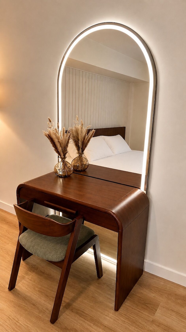 A wooden vanity with a mirror, a chair, and a bedside table with a lamp.