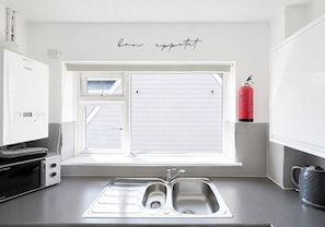 A kitchen with a double sink, a window with blinds, a white refrigerator, and a red fire extinguisher.