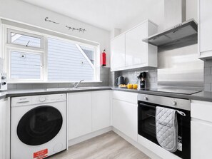 Una cocina moderna con muebles blancos, lavadora integrada, estufa y ventana con persianas.
