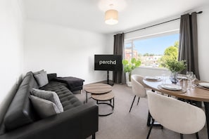 Una sala de estar moderna con un sofá de cuero negro, una mesita redonda y un comedor con una mesa de madera dispuesta para cuatro personas. Un televisor de pantalla plana está montado en la pared.