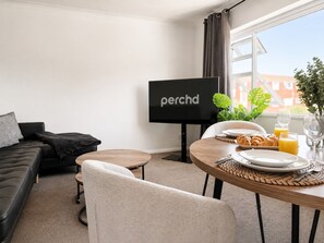 A modern hotel room with a black leather sofa, a round dining table with breakfast items, a flat-screen TV, and a view of buildings through the window.