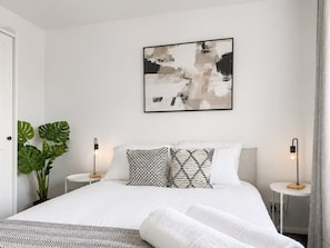 A modern bedroom with a large bed, two bedside tables, a wall-mounted abstract painting, and a potted plant.